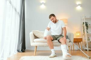 man recovery from accident fracture broken bone injury with leg splints in cast neck splints collar arm splints sling support arm in living room. Social security and health insurance concept photo