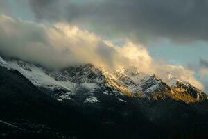 ai generado majestuoso montañas tapado con nieve, besado por el puesta de sol brillo. oscuro nubes telar, aún belleza prevalece. foto