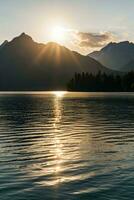 AI generated a tranquil scene of a sunset over a lake surrounded by mountains. The sun is partially obscured by the peaks of the mountains, casting long rays of light across the sky. photo