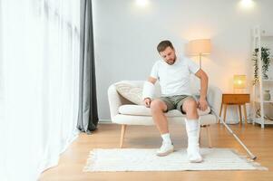 man recovery from accident fracture broken bone injury with leg splints in cast neck splints collar arm splints sling support arm in living room. Social security and health insurance concept photo