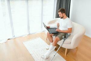 Happy young man with arm in a cast sitting on the couch at home and communicating on a laptop photo