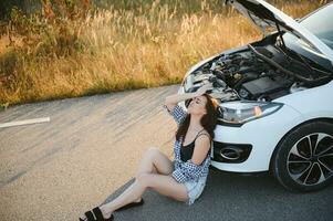 A young girl sits near a broken car on the road with an open hood. photo