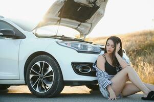 Beautiful young girl sitting at a broken car on the road desperate to get help photo