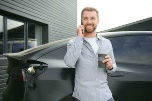 handsome bearded man in casual wear, standing at the charging station and charger for an electric car. Eco electric car concept photo