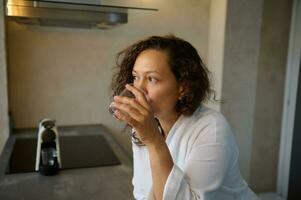 Close-up portrait of a Latin American young adult confident pretty woman in white robe, drinking water in the morning. photo