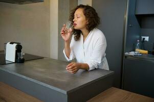 Young adult Hispanic woman in a white robe taking medication, drinking water, standing at the kitchen counter photo