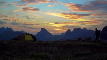 uma turista com uma mochila goza uma lindo nascer do sol perto uma barraca dentro a montanhas. lindo Visão do a atingiu o pico tops do a lofoten ilhas. Noruega 4k video