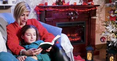 mãe lendo uma história para dela filha a partir de uma livro em Natal dia com Natal decoração dentro a casa. video
