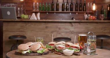 Very big selection of burgers on wooden table in front of a bar. A plate full of burgers of different type on a vintage table in a pub. video