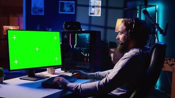 Man in dimly lit apartment playing video games on isolated screen gaming PC at computer desk, enjoying day off from work. Gamer battling enemies in online multiplayer shooter on chroma key monitor