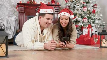 Smiling couple sitting on the floor shopping online using a smartphone in Christmas decorated room video