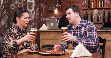 Couple discussing while drinks beer and eats pizza in very cozy and vintage pub or restaurant. There is a bearded hipster barman in the background working at the counter. video