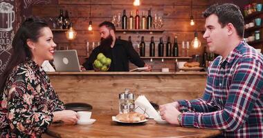 mooi Kaukasisch paar aan het eten croissants en drinken koffie in knus vestiging. in de achtergrond - een gebaard barman is werken Bij de teller video