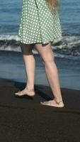 Waist down view of woman barefoot walking along beach of black sand of Pacific Ocean. Vertical video