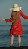 Rear view unrecognizable woman in summer red polka dot dress dances, standing on black sand of beach video