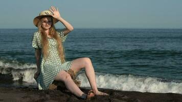 Young blonde in summer straw hat polka dots dress sits on log on shore of sea with black sand beach video