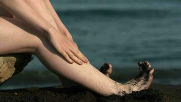Closeup of barefoot woman shakes feet off black sand on beach on background of ocean waves video