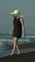 Barefoot mature adult woman walking on black volcanic sand of Pacific Ocean beach. Vertical video