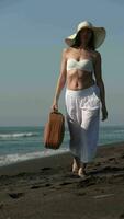 maduro mulher caminhando com Castanho mala de viagem em Preto arenoso de praia em pacífico oceano durante de praia feriados video