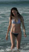 caucásico mujer en bikini caminando en playa rodilla profundo en agua de rotura olas de Pacífico Oceano video