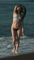 lleno longitud mujer en bikini caminando negro arenoso playa tobillo profundo en agua de olas de Pacífico Oceano video