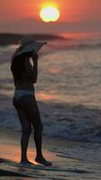 mujer en blanco bikini, Paja Dom sombrero caminando en playa a amanecer. hembra tobillo profundo en rotura ola video