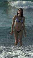 frente ver Delgado 50 años antiguo mujer en bikini caminando en playa rodilla profundo en agua de rotura olas video