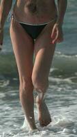 irreconhecível mulher com grandes pernas dentro bikini inferior caminhando em de praia dentro espirrando quebra ondas video
