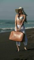 ensam 50-åring kvinna med brun resväska stående på svart sandig strand och ser bort video