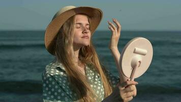 Woman holding mirror with hand and looking into it, straightens her long hair on background of sea video
