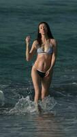 Full length slim woman in bikini walks on sandy beach ankle deep in water of waves of Pacific Ocean video