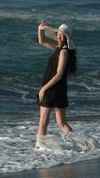 Woman in short summer dress and straw hat standing in sea waves, turning around, looking at camera video