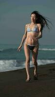 frente ver de contento morena mujer en bikini corriendo en negro arenoso playa de Pacífico Oceano video