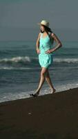 lleno longitud descalzo mujer caminando en negro arenoso playa. caucásico hembra en turquesa verano vestir video