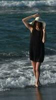 Playful barefoot woman in breaking waves on black sandy beach. Female in summer dress and straw hat video