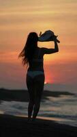 Woman in white bikini walking on sandy beach at sunrise, putting on straw hat during summer vacation video