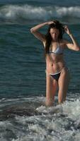 full längd av 50 år gammal kvinna i bikini gående på strand i vatten av stänk brytning vågor video