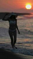 Woman in white bikini, straw sun hat walking on beach at sunrise raises hand and shows victory sign video