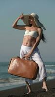 50-year-old Caucasian female holding brown suitcase posing on black sandy beach, looking away video