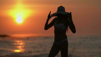 kvinna i bikini, sugrör Sol hatt på strand på soluppgång. kvinna silhuett på bakgrund stor Sol på gryning video