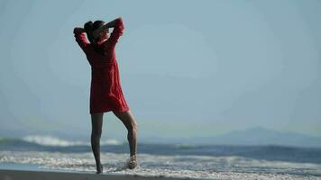 Serene woman stands ankle deep in water breaking wave on sandy beach, hands behind head. Slow motion video