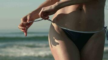Closeup woman in bikini bottom tying straps on panties, standing on background breaking wave video