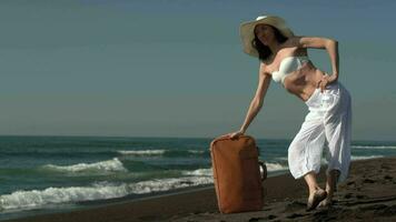 Confident woman standing on sandy beach leaning on suitcases, looking away at waves of Pacific Ocean video