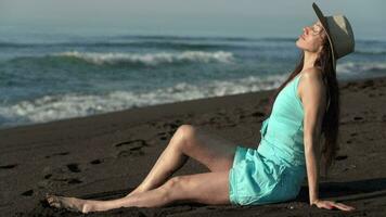 50-year-old barefoot woman sitting on beach, sunbathing relaxed in short summer dress, straw sun hat video