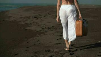 Cropped waist down back view of unrecognizable woman walking on beach holding brown suitcase video