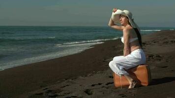 mujer sentado en maleta en negro arenoso playa, mirando lejos a hermosa calma olas Pacífico Oceano video