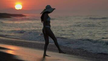 Woman tourist in white bikini and straw sun hat walking on sandy beach at sunrise on summer holiday video