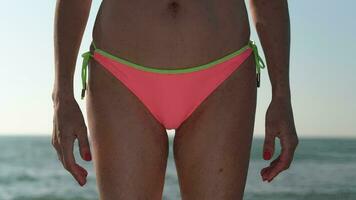 Closeup front view of slim woman in pink bikini bottom standing on background breaking wave of ocean video