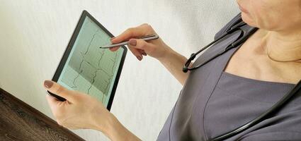 Shot of the female doctor in uniform against the white wall studying cardiogram on the tablet. Healthcare photo