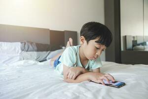 Close up portrait of Asian boy with black bangs, black eyes with a smiling face wearing a light green shirt lying on the bed in his house playing with his smart phone. Education concept. photo